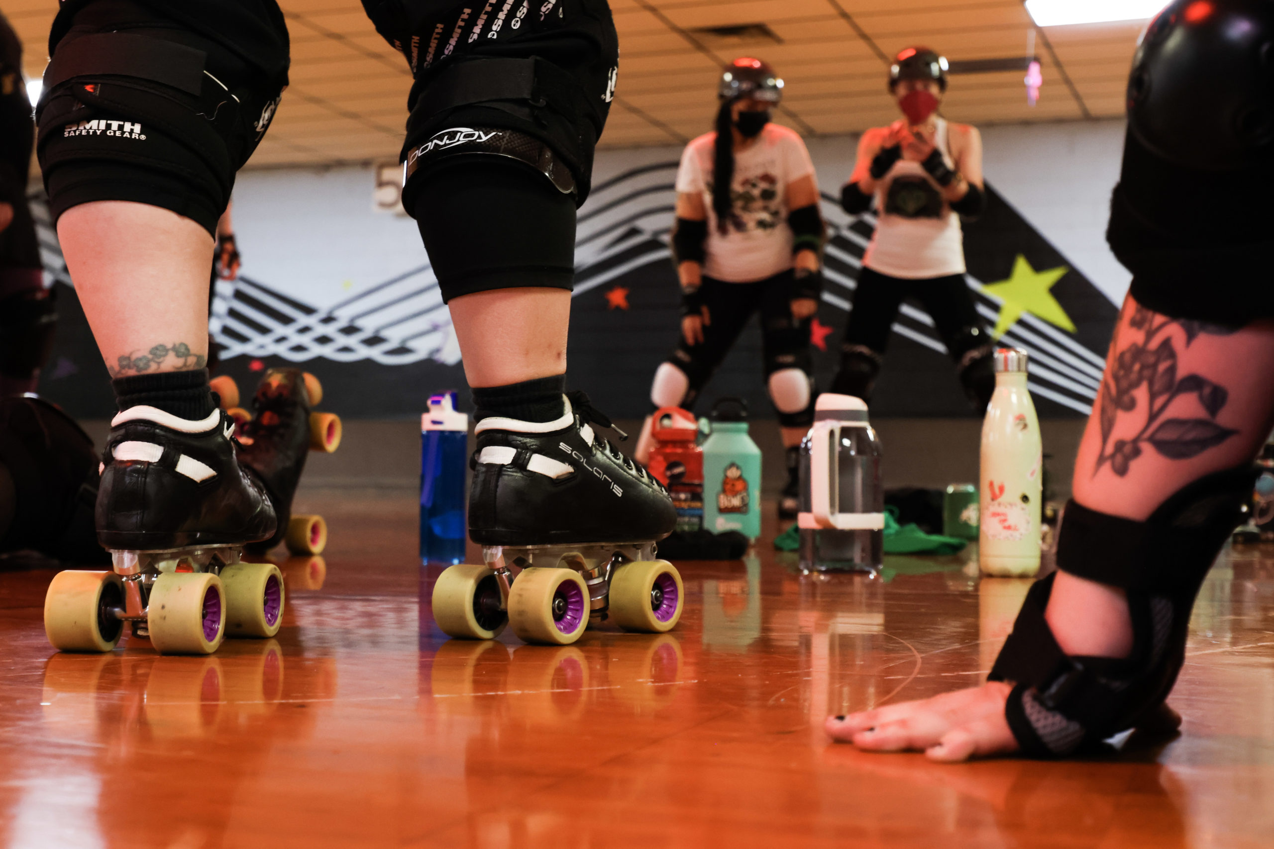 A close up photo of a roller derby skater’s roller skates and another skater’s hands. Both skaters watch coaches in the background explaining a drill at roller derby practice.