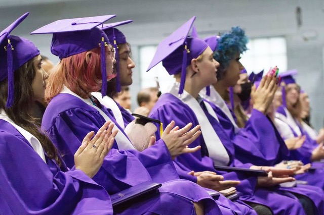 A row of students wearing purple gowns sit at a graduation ceremony.