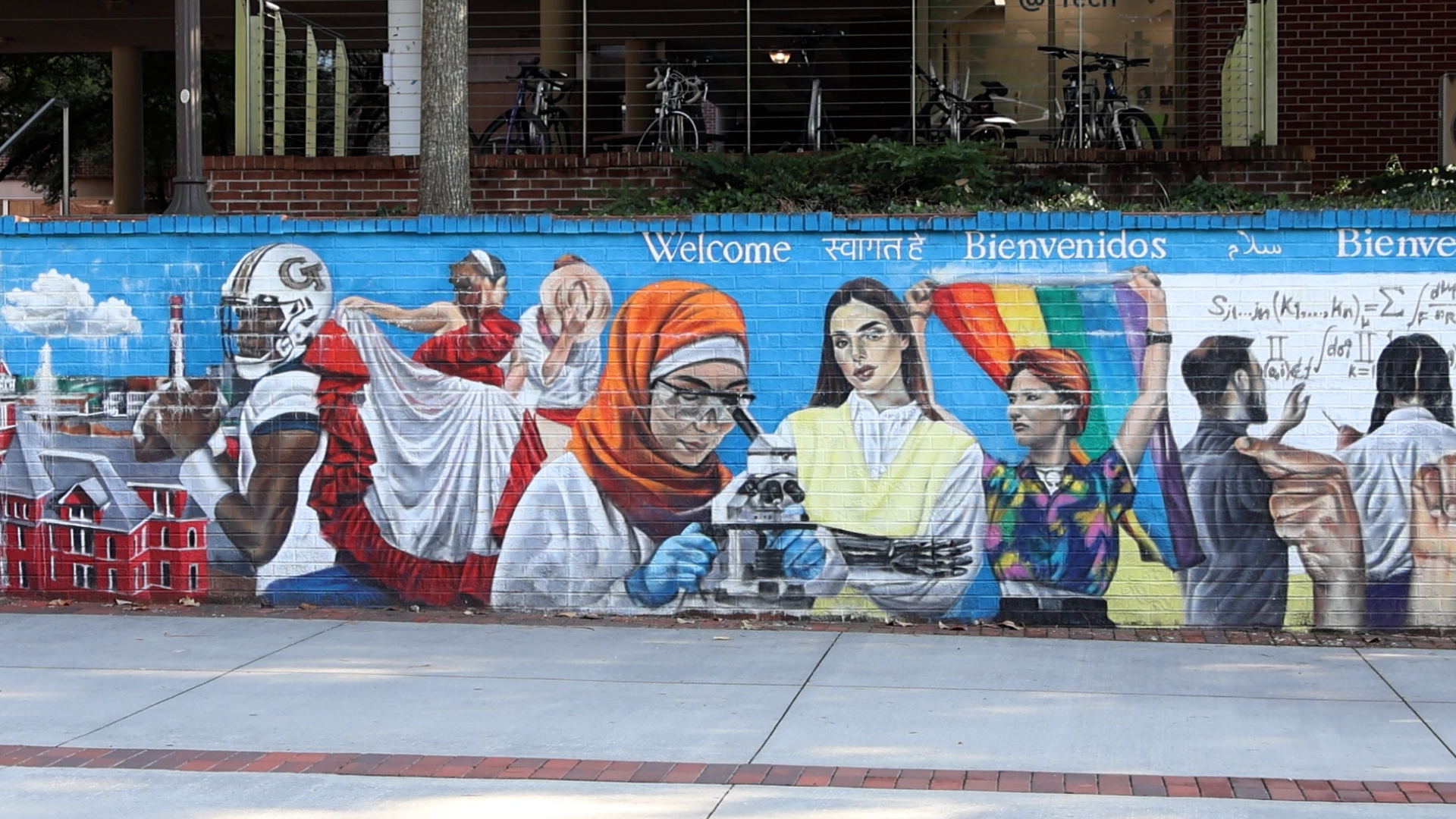 A mural on Georgia Tech’s campus shows a diverse student body including a Black football player, a flamenco dancer, a hijabi scientist, an amputee, and a person holding a pride flag. Above the students are words for “welcome” in different languages.